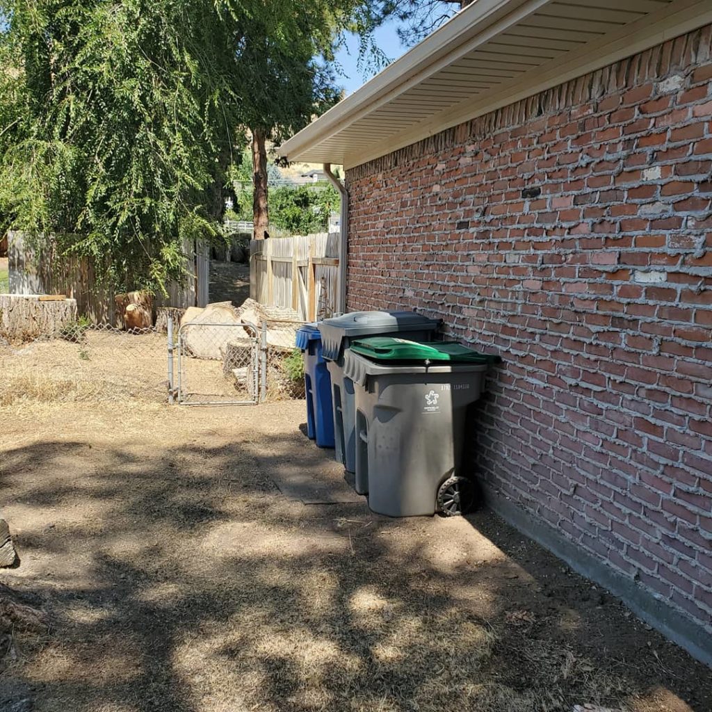 Image of trash containers by brick wall on side of house where logs have been cleared